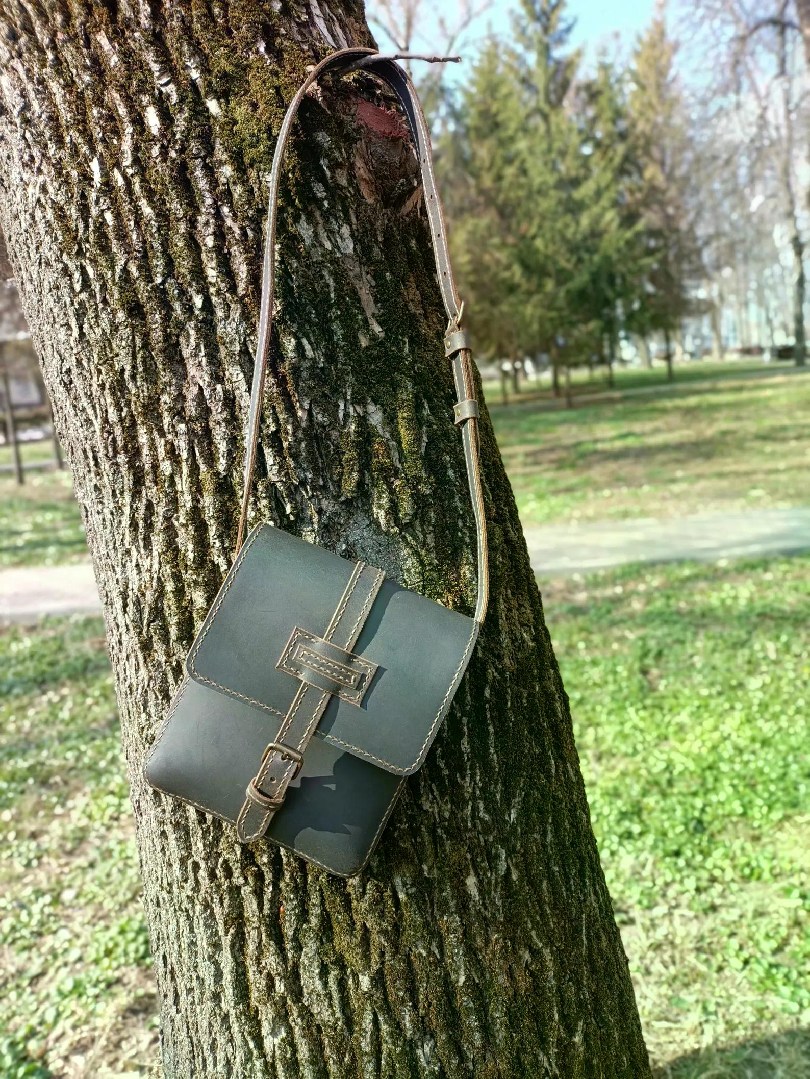 Handmade gray leather crossbody messenger bag hanging from a tree outdoors